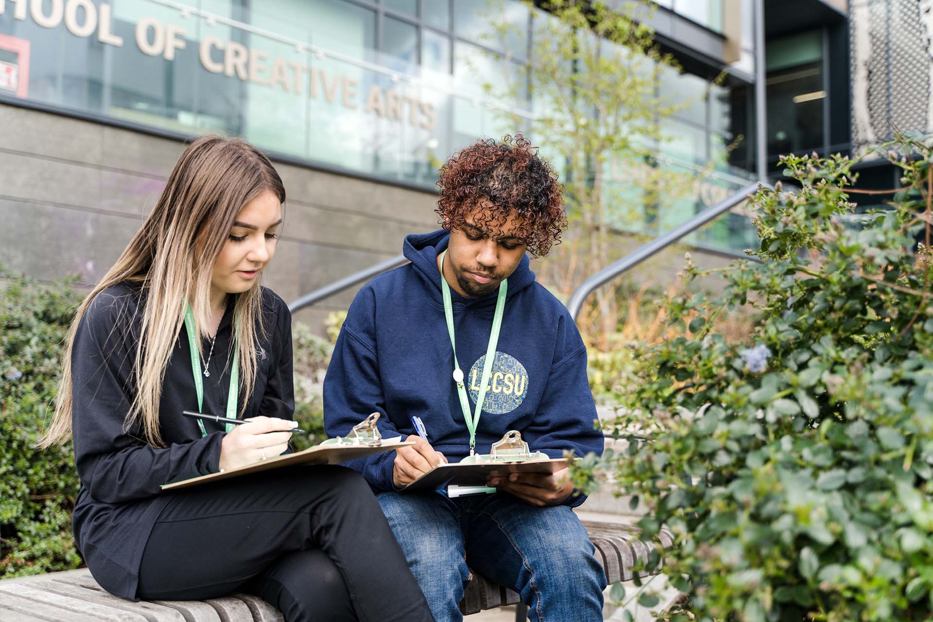Two students sat outside Quarry Hill campus, looking down at clipboards holding pens, wearing college lanyards