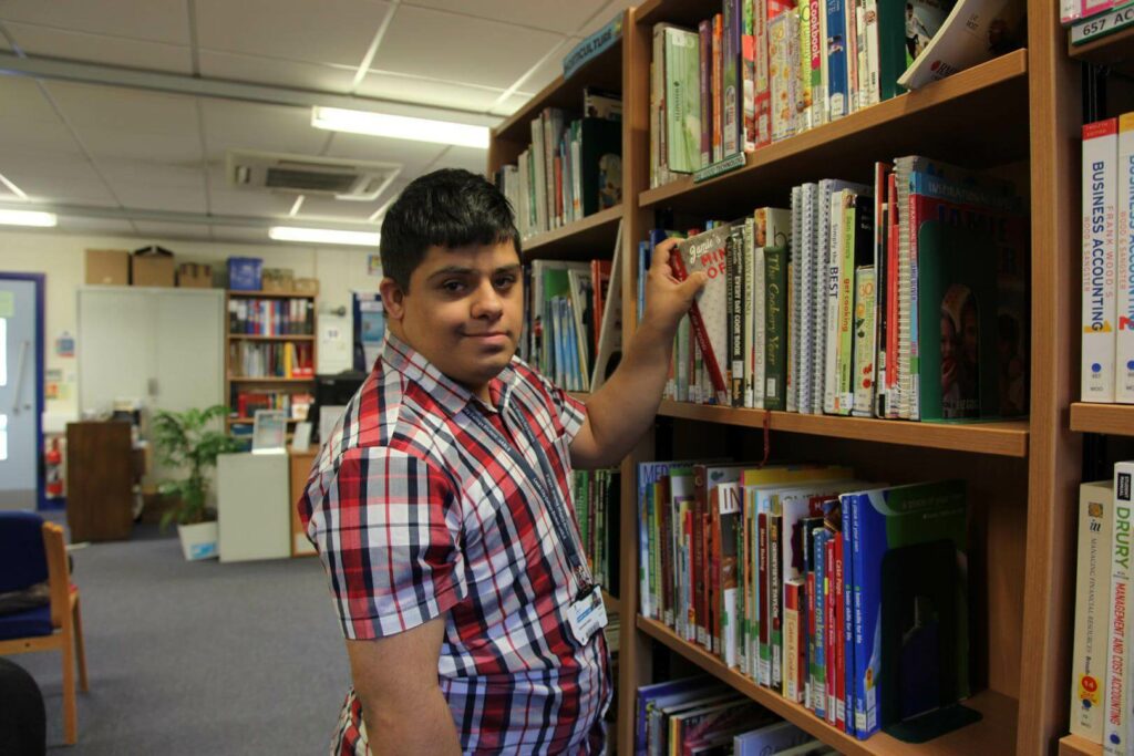 Student in library taking a book off the shelf smiling at the camera