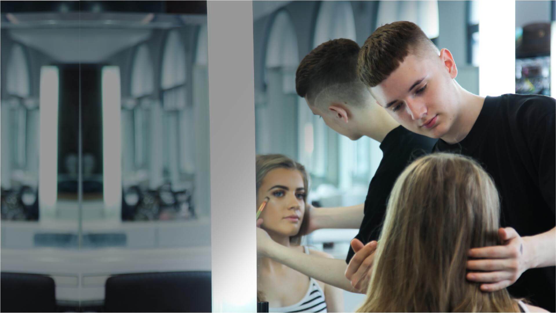Student applying make up to someone's cheek with a makeup brush whilst they are sat in front of mirrors in the makeup studio