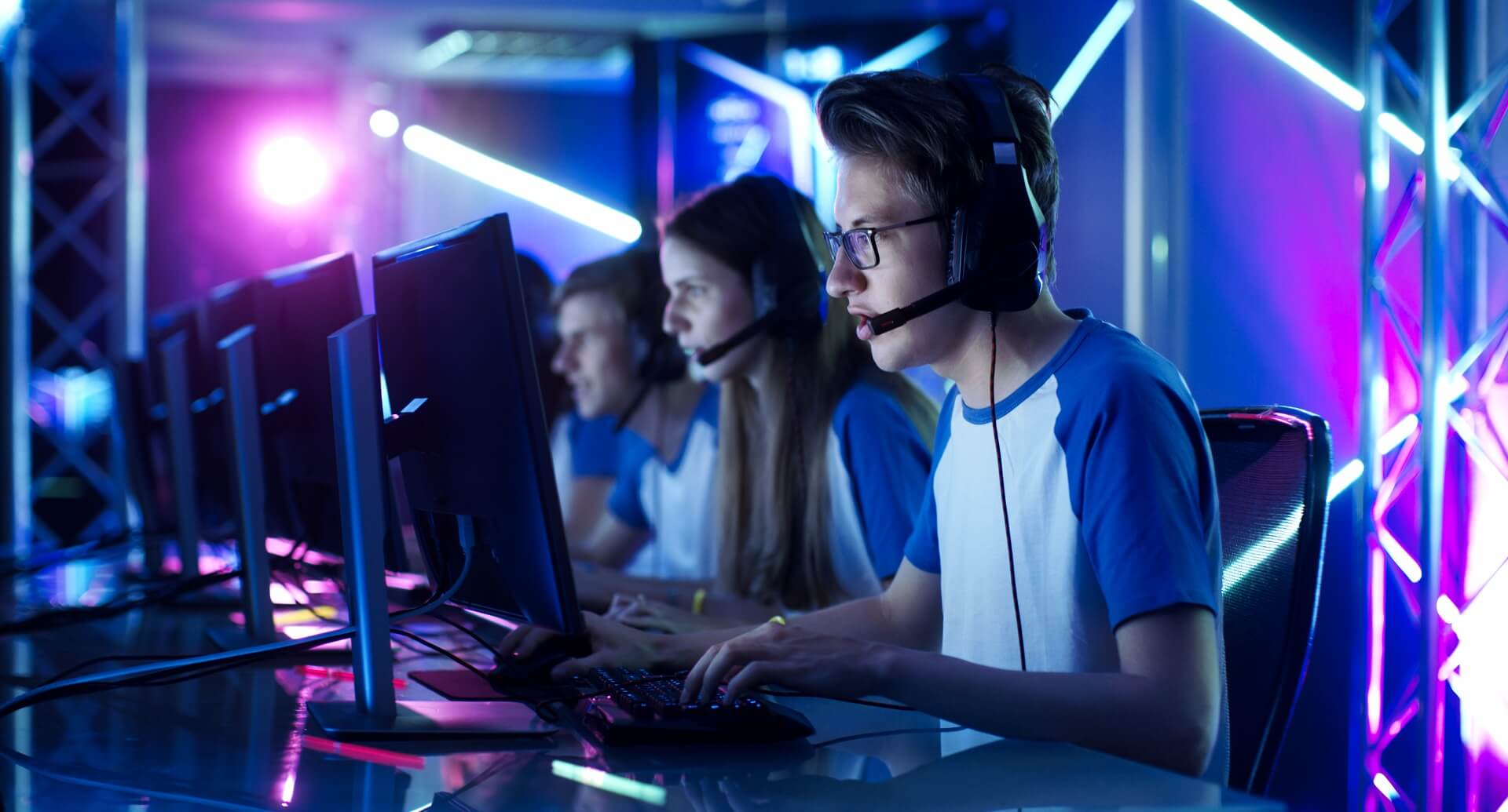 People sat in front of computer screens wearing headsets in a dark room with neon lighting