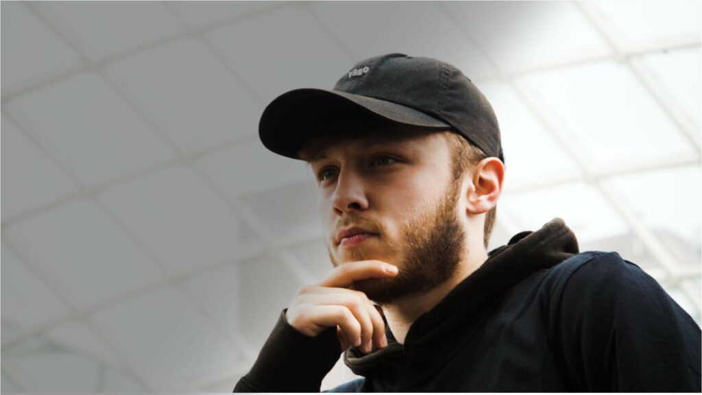 Student wearing a black hoodie and a cap rubbing chin looking pensive