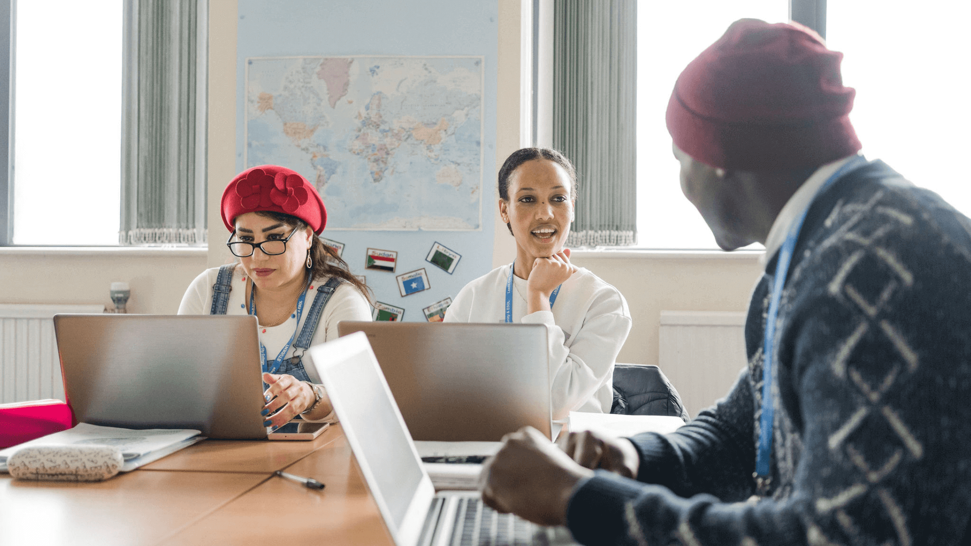 Three adult students sat in a classroom looking at laptops on the desks and chatting