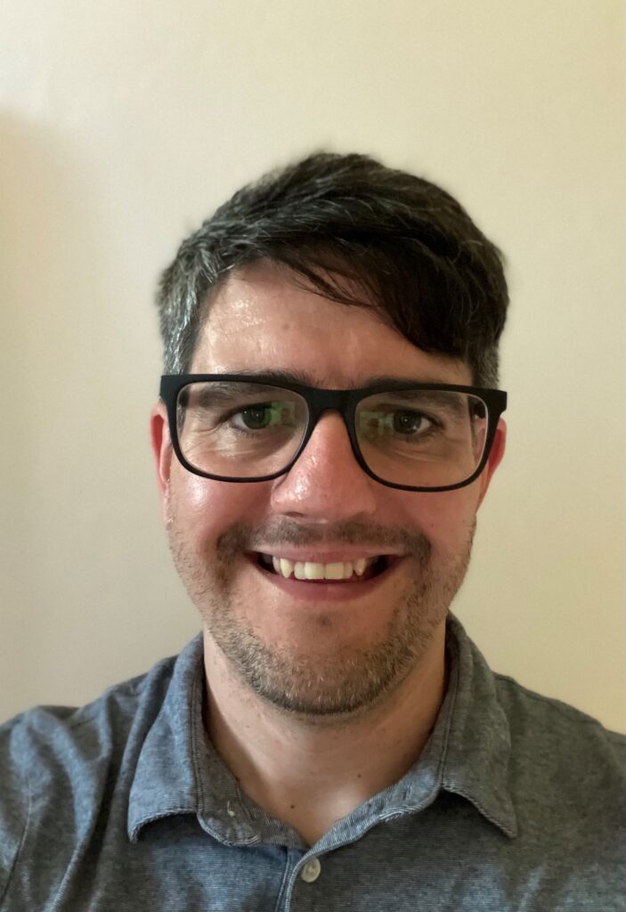 Close up selfie of a person smiling warmly, wearing glasses and wearing a button up shirt