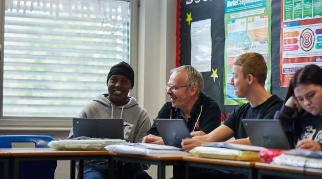 14-16 students and teacher sat in classroom working on laptops and smiling