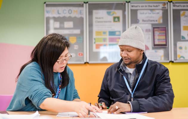 Leeds City College adult student sat with teacher in classroom looking at work together on the desk, both wearing Leeds City College lanyards