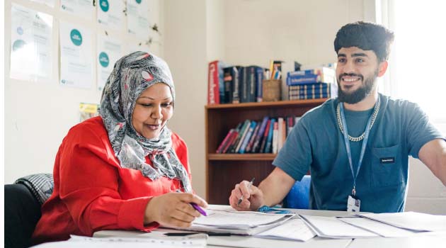 Two adult students sat at a desk in a classroom looking at a workbook together and smiling