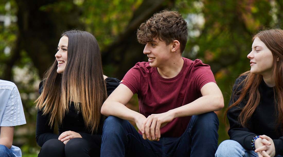 Group of students sat on a wall outside with trees behind, looking to the side and smiling