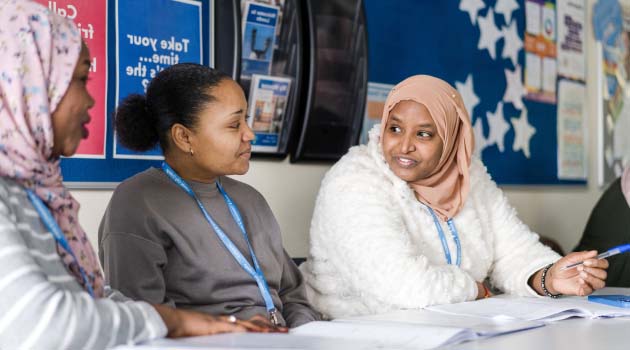 Three Leeds City College adult students wearing blue lanyards sat at a table in a classroom smiling and discussing work in the textbooks in front of them
