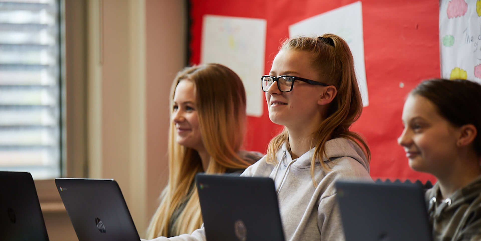 14+ Academies students sat by laptops in classroom looking up smiling