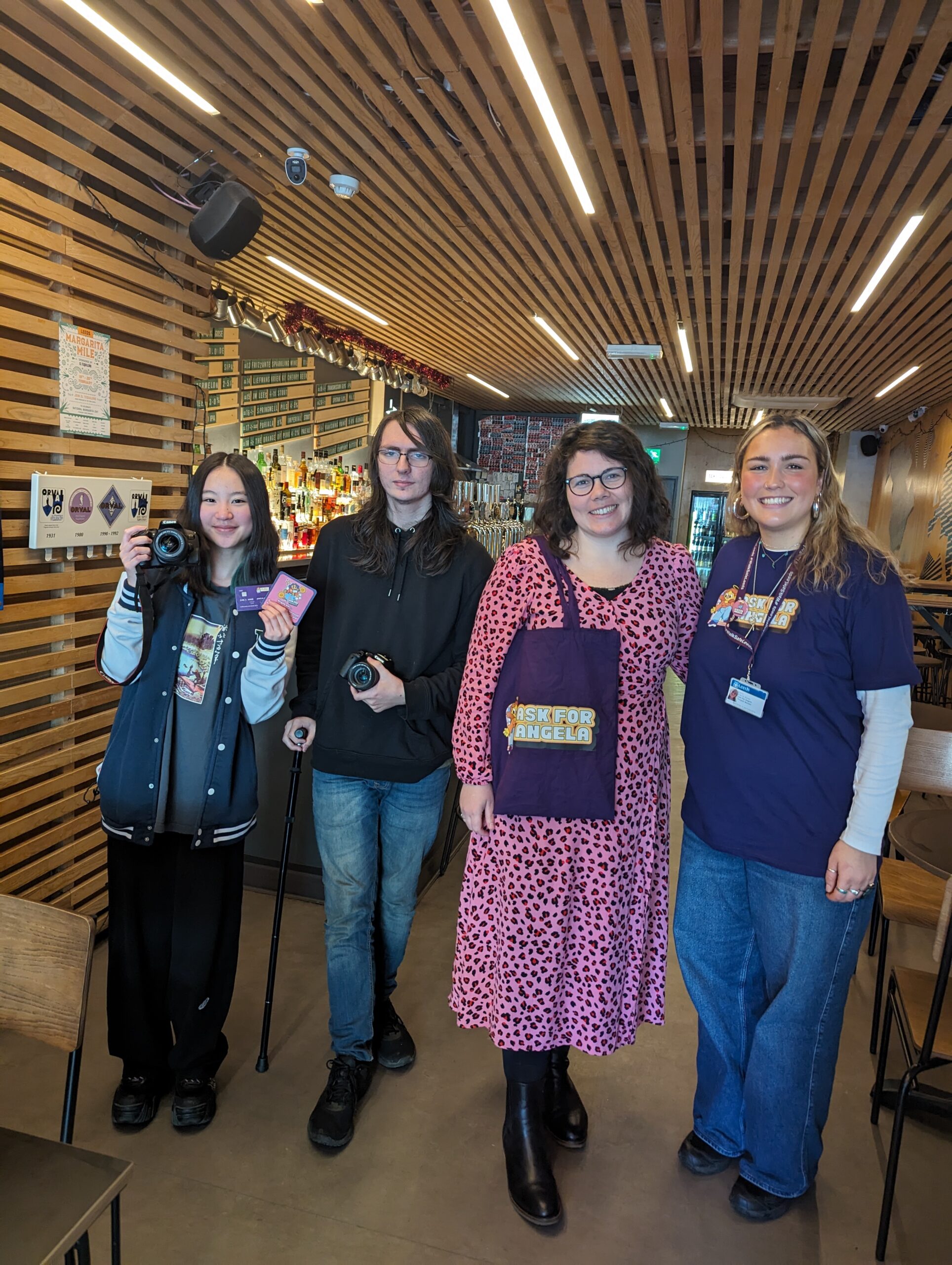 Four people stand in a cafe smiling at the camera, two holding film equipment, one holding an 'Ask Angela' bag