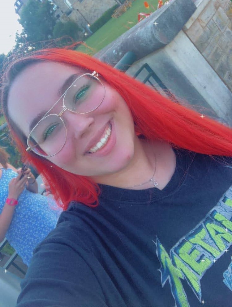 A woman with vibrant red-coloured hair smiles at the camera whilst standing outside