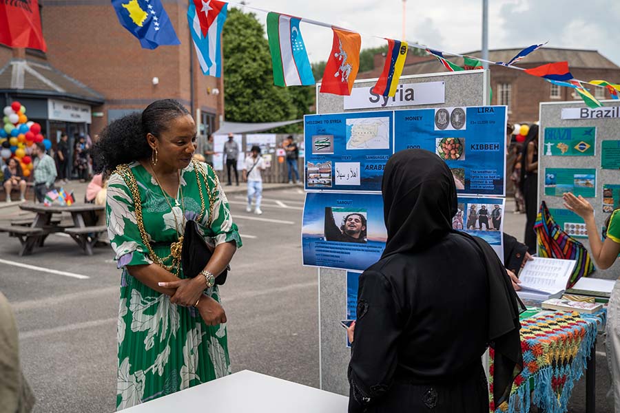 Two people at the International Day 2024 looking at boards with information on Syria and Brazil, with international flag bunting and balloon decorations on the street