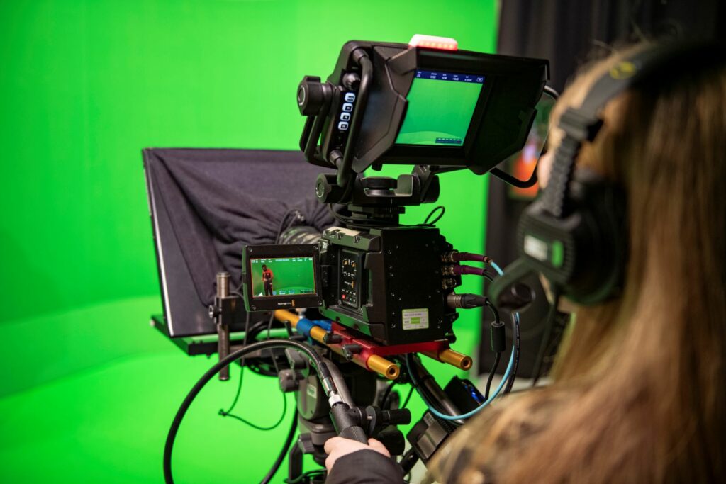 A studio camera being operated to film a student in a green screen room