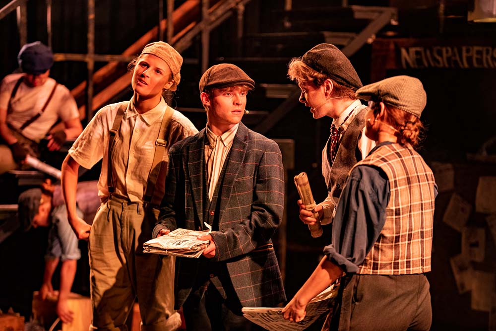 Group of four Leeds City College Creative Arts Performing Arts students acting on stage wearing flat caps, waistcoats and holding newspapers
