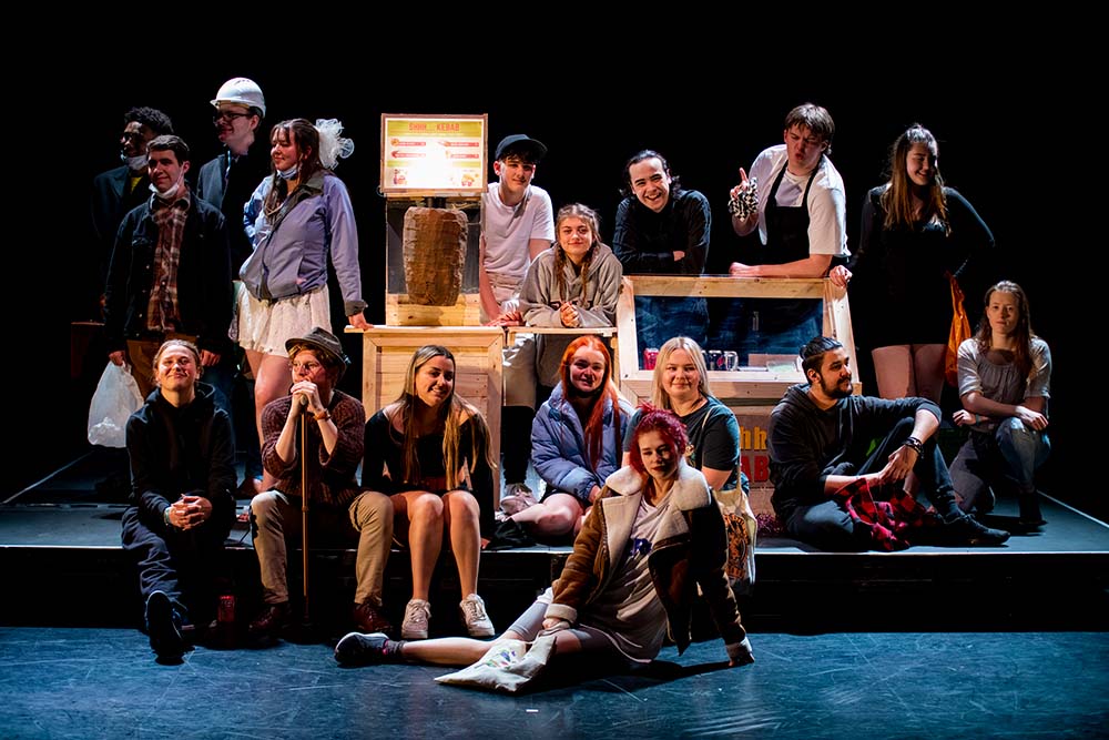 Group of Leeds City College Creative Arts students performing on stage in a production of the Kebab Chronicles, all sat around a kebab