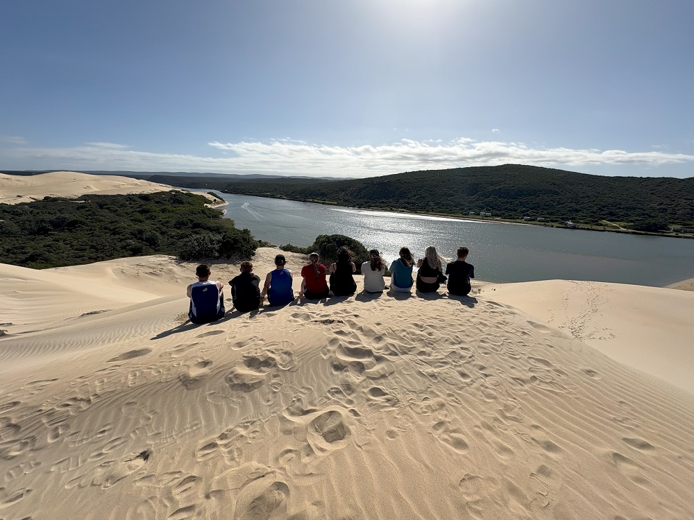 Coaching students from Leeds City College sitting on a sand dune overlooking water and hills in South Africa