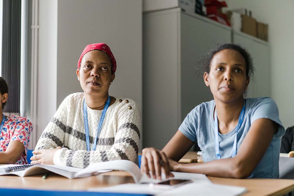 Two adult students sat at desks in a classroom at Deacon House wearing Leeds City College lanyards looking up